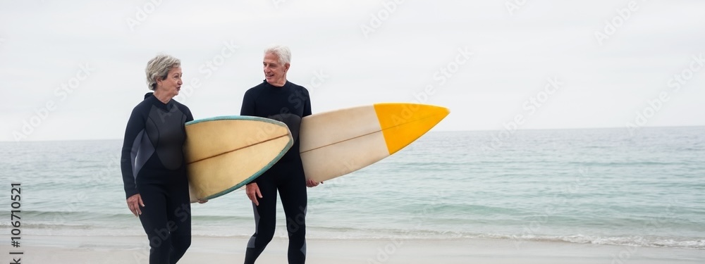 Older couple in wetsuits carrying a surfboard on the beach